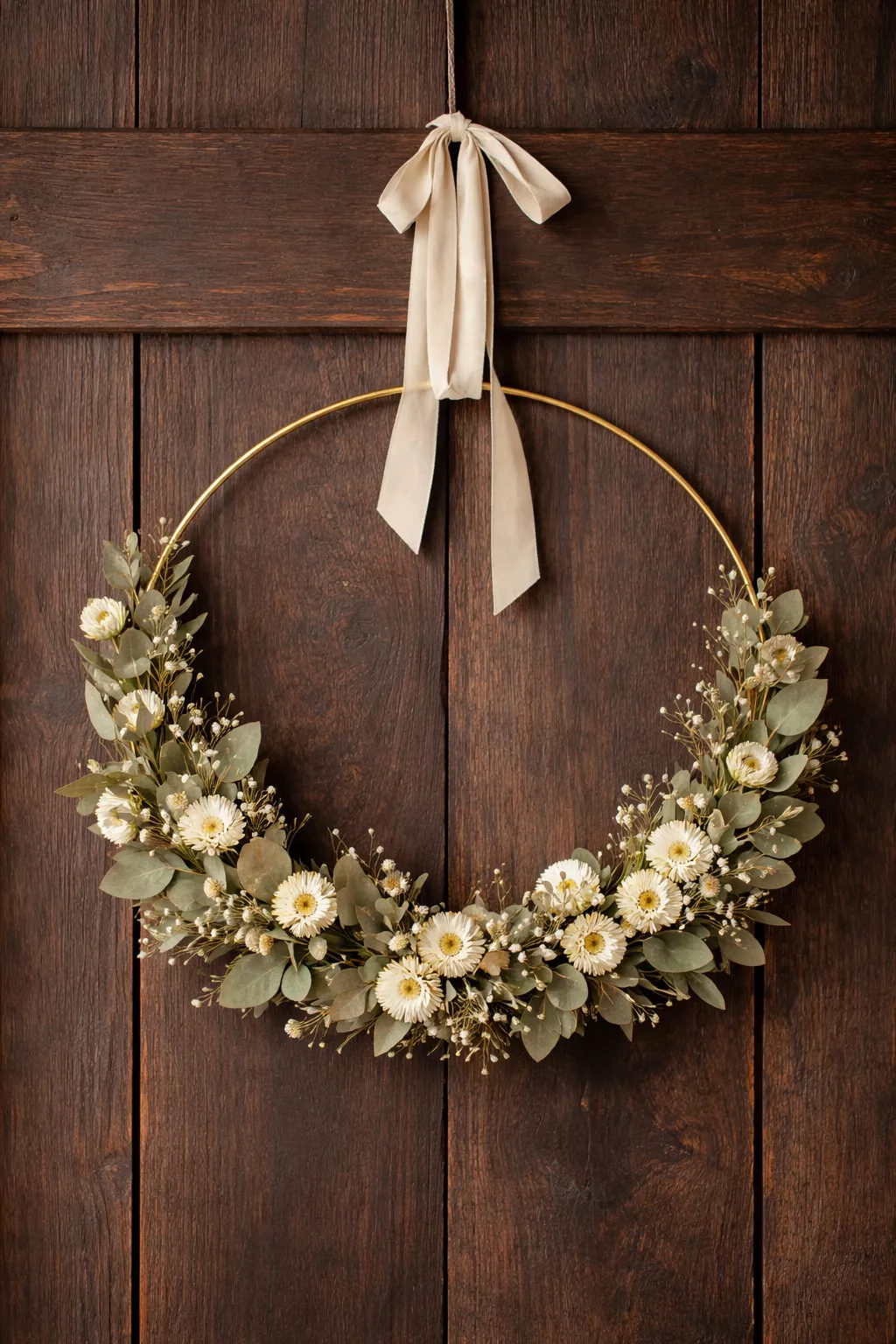 A realistic photo of a circular gold metal hoop wreath adorned with dried eucalyptus and white everlastings, hanging on a rustic dark wood door with a cream velvet ribbon.