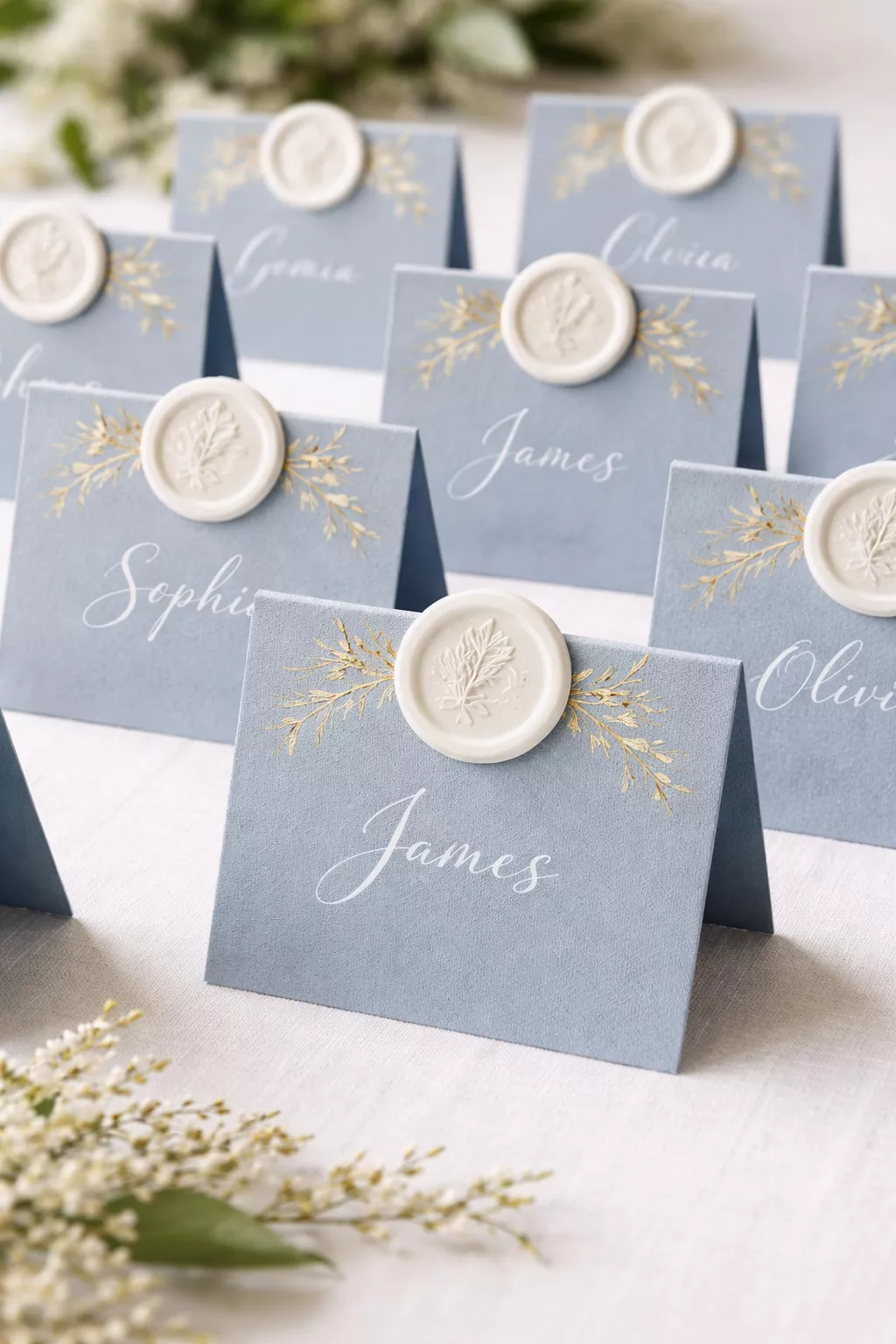 A realistic photo of dusty blue cardstock place cards with white wax seals and gold foil botanical motifs on a white tablecloth