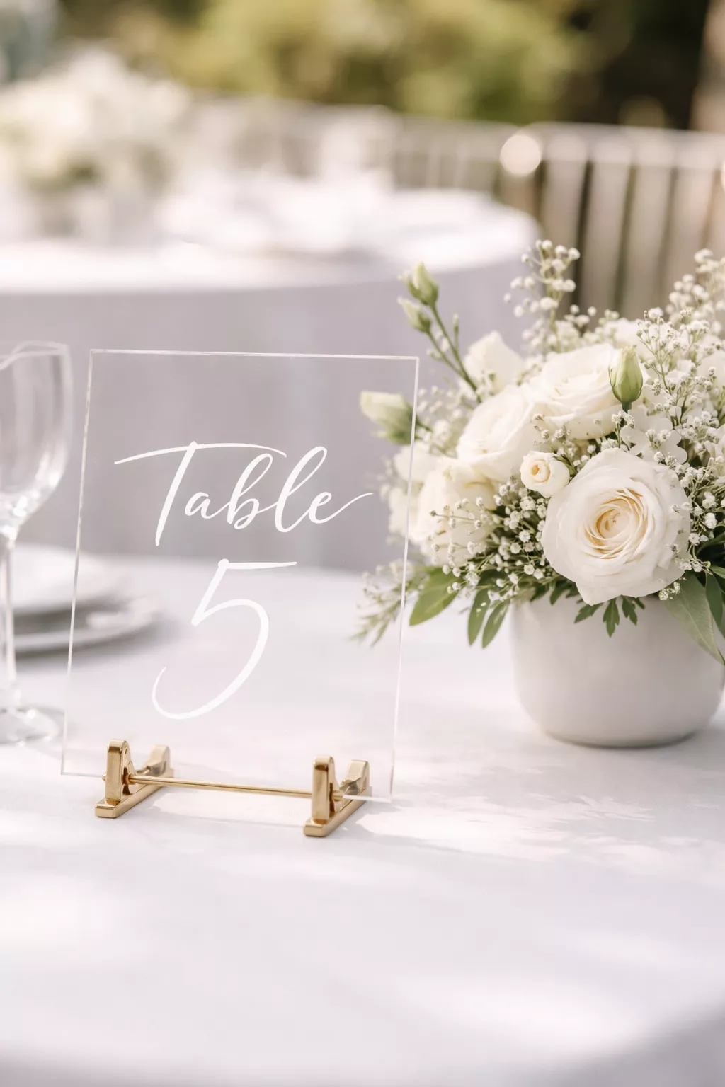 A realistic photo of a clear acrylic table number with elegant white hand-lettered calligraphy, propped up on a small gold easel next to a small white floral arrangement.