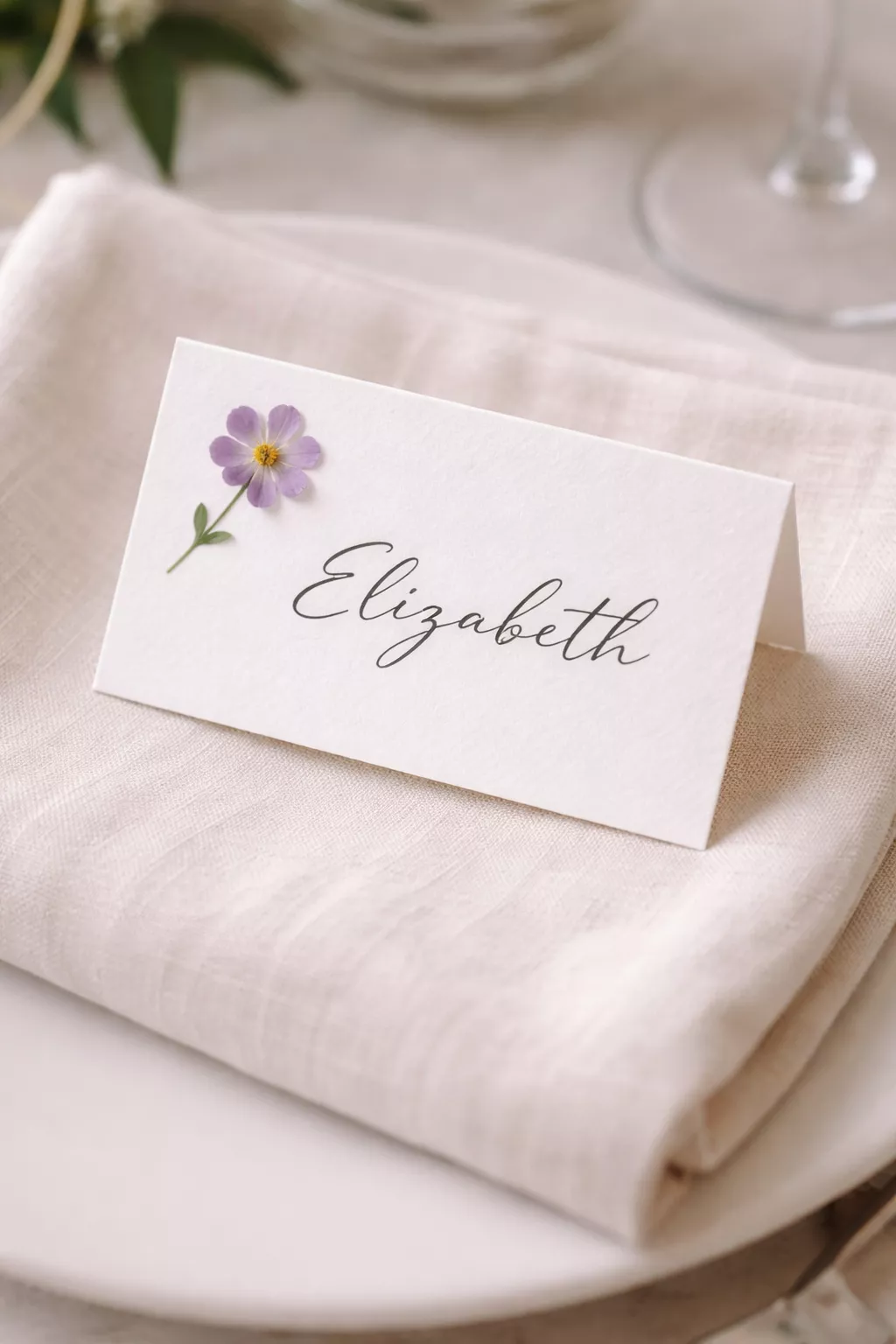 A realistic photo of a white wedding place card featuring a single small purple pressed flower on a linen napkin.