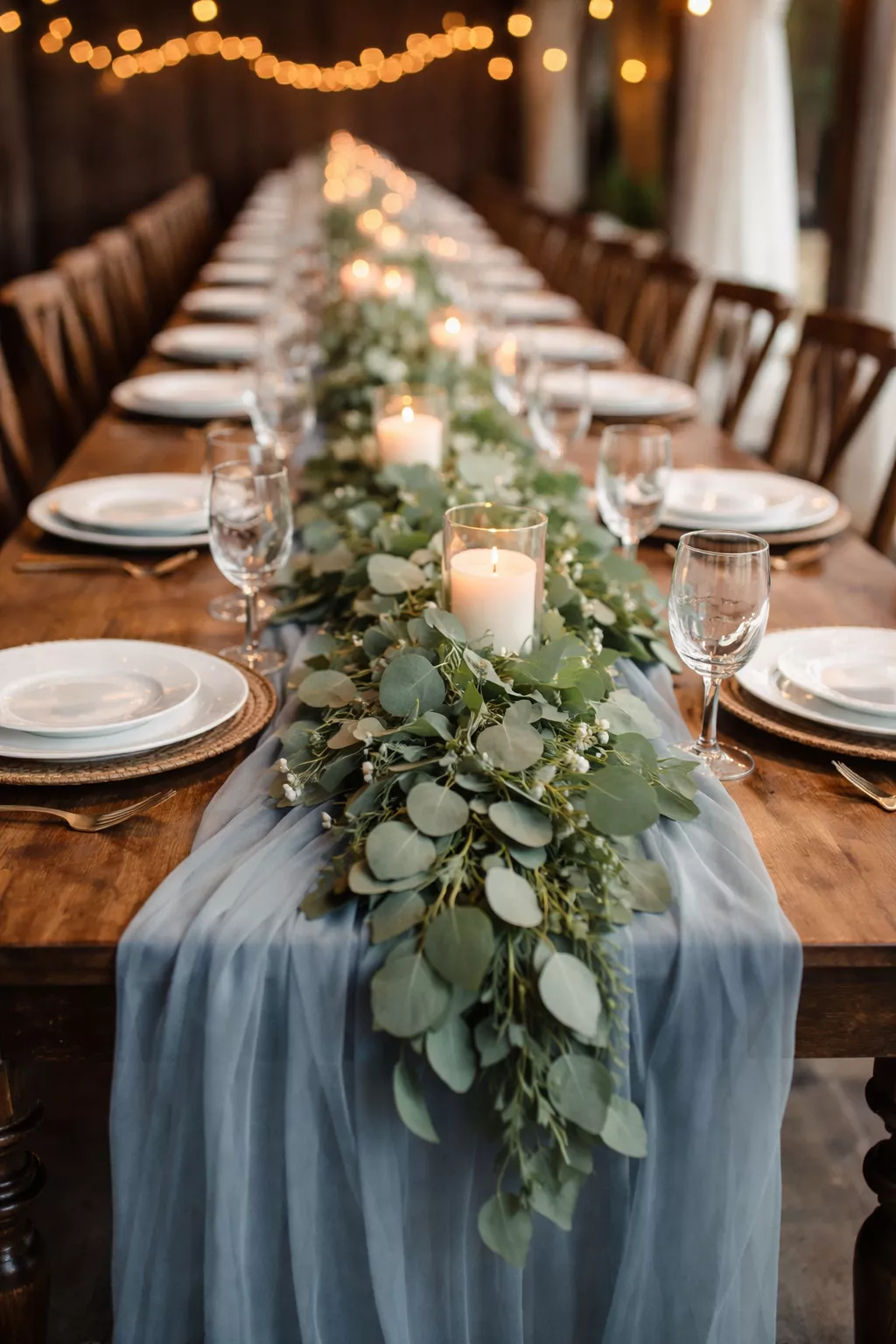 A realistic photo of a long wooden dining table with a dusty blue chiffon runner and sage green eucalyptus garland under soft lighting