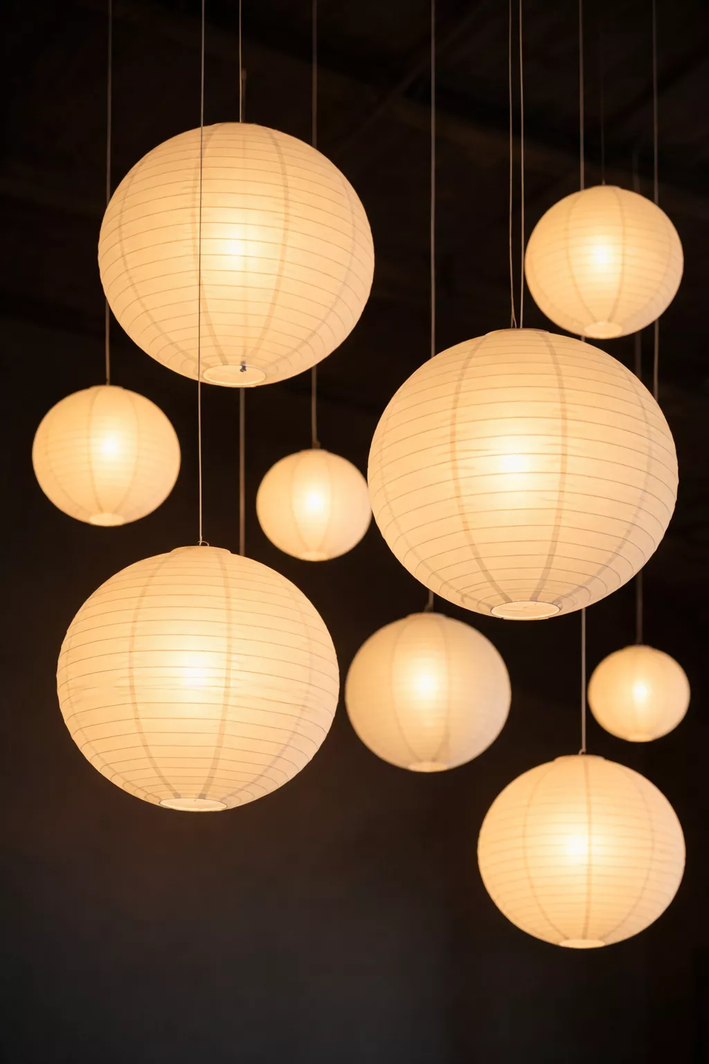 A realistic photo of several round white paper lanterns of various sizes hanging from a dark ceiling with thin fishing line, glowing with warm internal light.