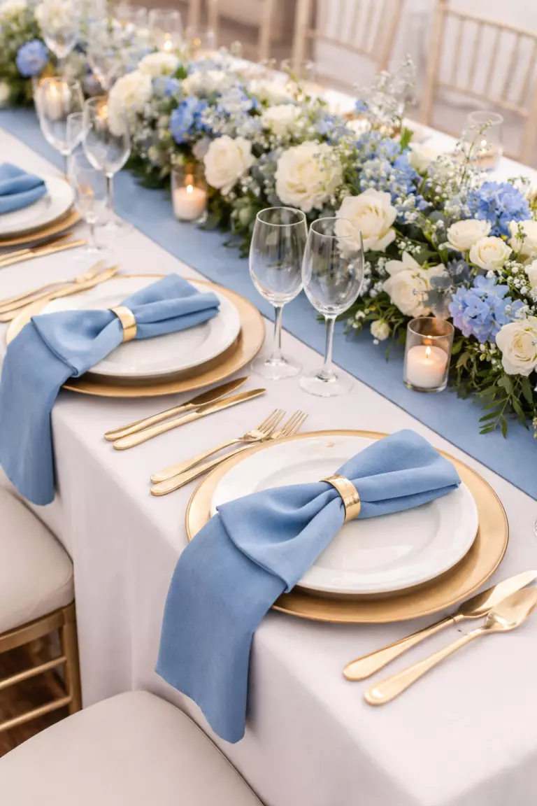 A realistic photo of a wedding table setting with a cornflower blue linen table runner, matching napkins, gold chargers, and white ceramic plates.