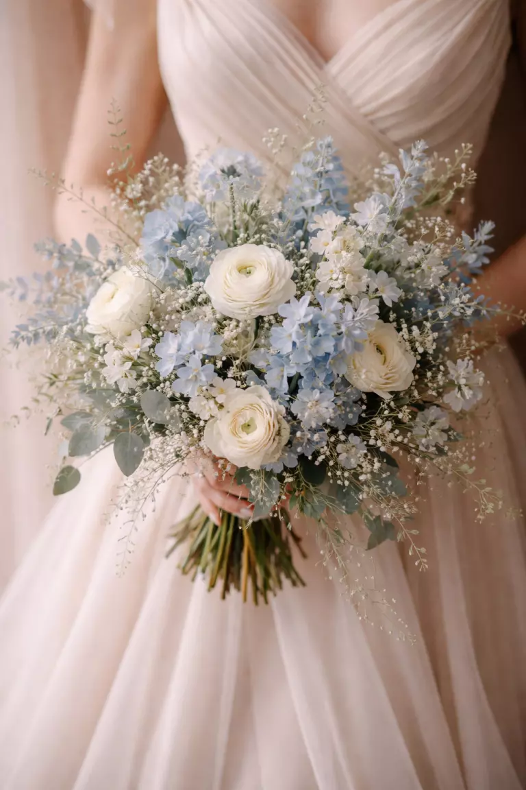 A realistic photo of a loose wedding bouquet with dusty blue wildflowers and cream blossoms, meadow style, soft and airy, held against a champagne colored dress.