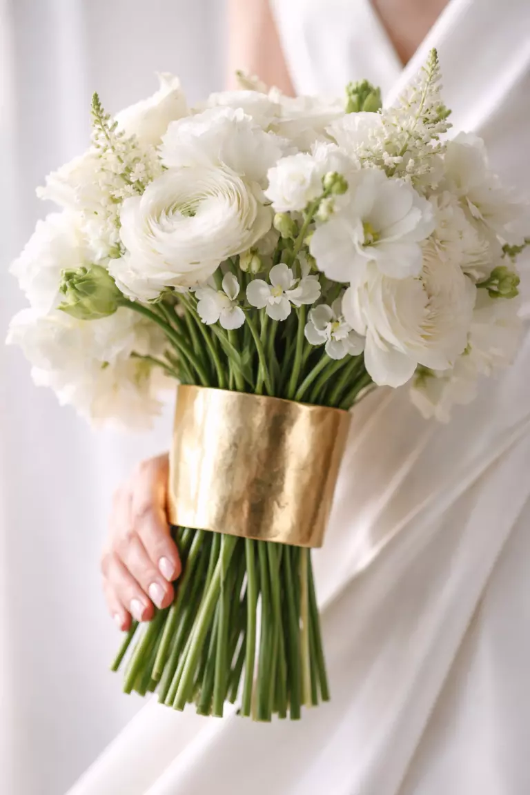 A realistic photo of wedding flower stems encased in a hand forged brass cuff, modern floral design with soft white petals, couture fashion style, bright lighting.
