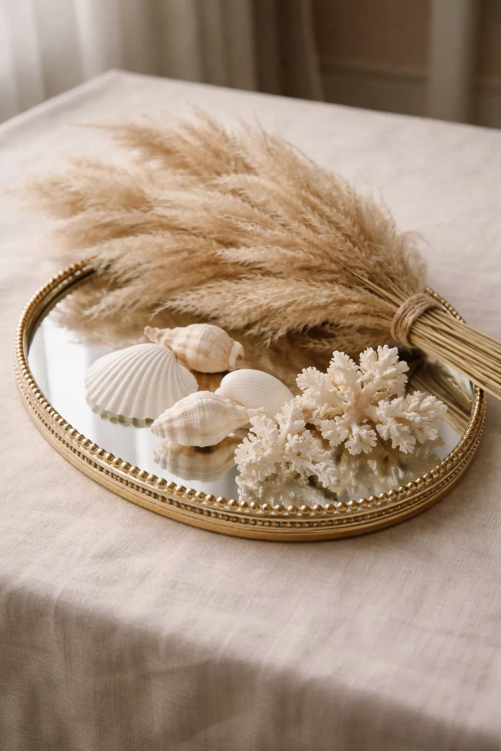 A realistic photo of a mirrored tray holding tan pampas grass, white shells, and small coral on a neutral linen tablecloth.