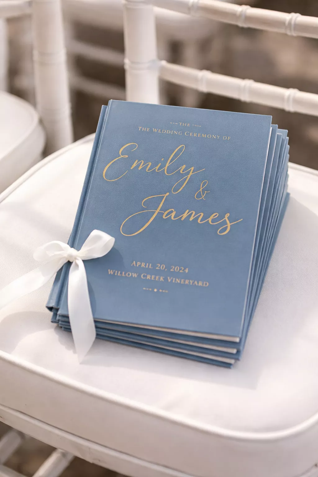 A realistic photo of wedding ceremony programs with dusty blue covers and gold foil lettering sitting on a white chair