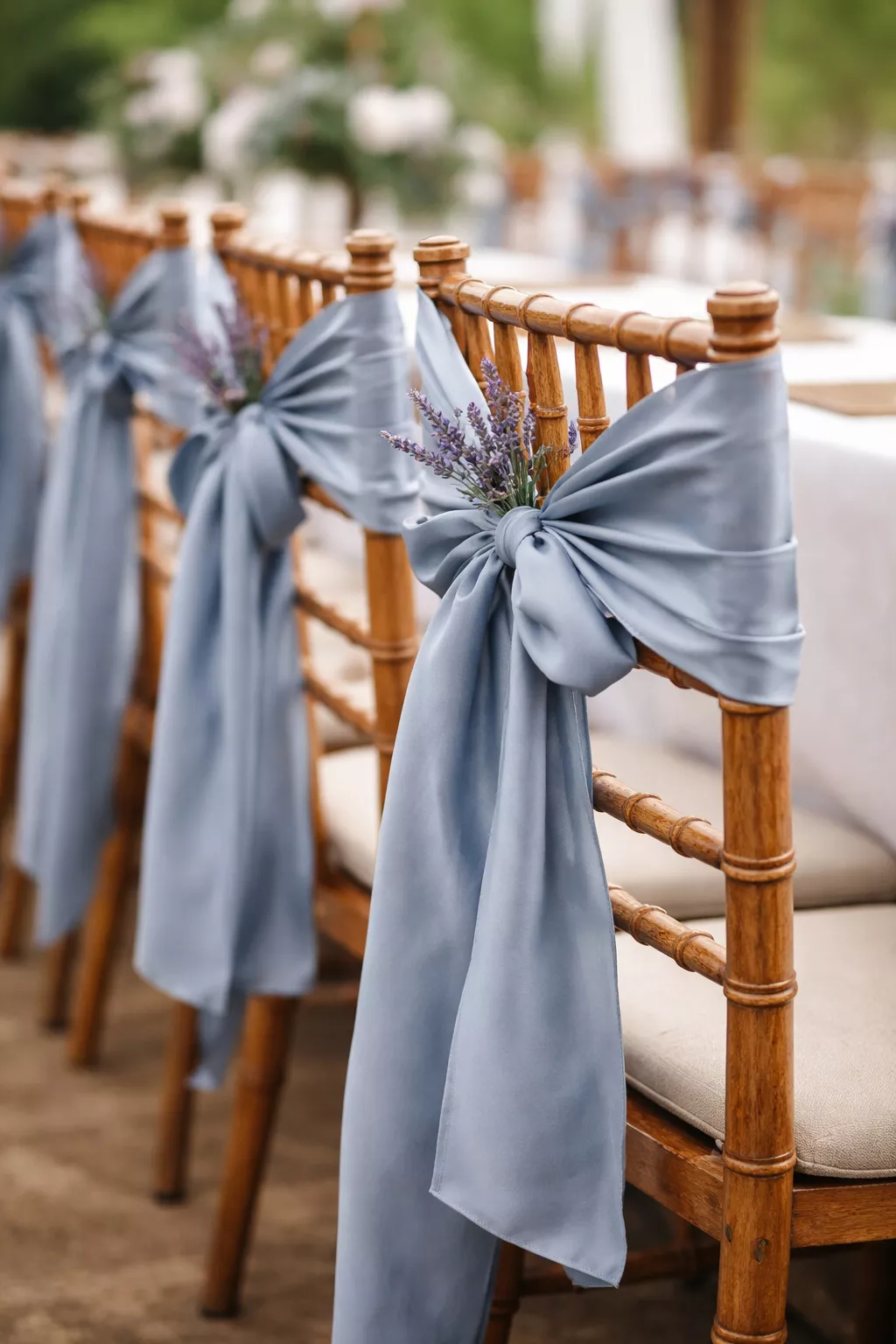 A realistic photo of wooden farm chairs with dusty blue satin sashes tied in elegant bows and small sprigs of lavender tucked into the knots