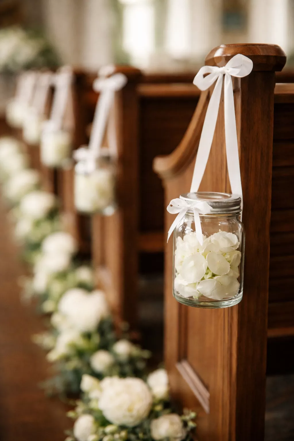 A realistic photo of small glass jars filled with white rose petals hanging from the end of wooden pews with simple white satin ribbons.