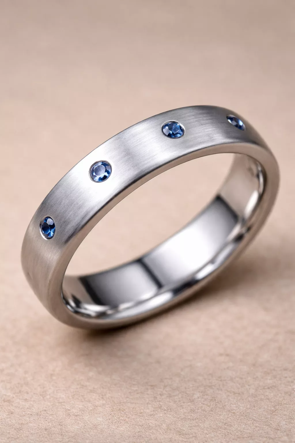 A realistic photo of a brushed silver wedding band with small blue sapphires set flush into the metal surface.