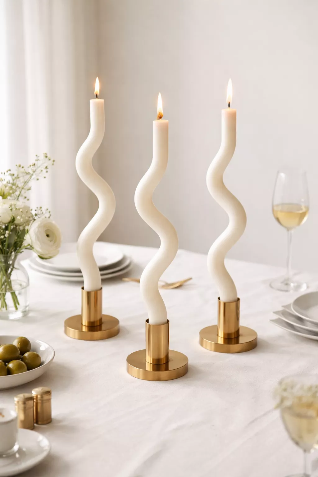 A realistic photo of long white taper candles that have been bent into wavy shapes, standing in modern gold candle holders on a white tablecloth.