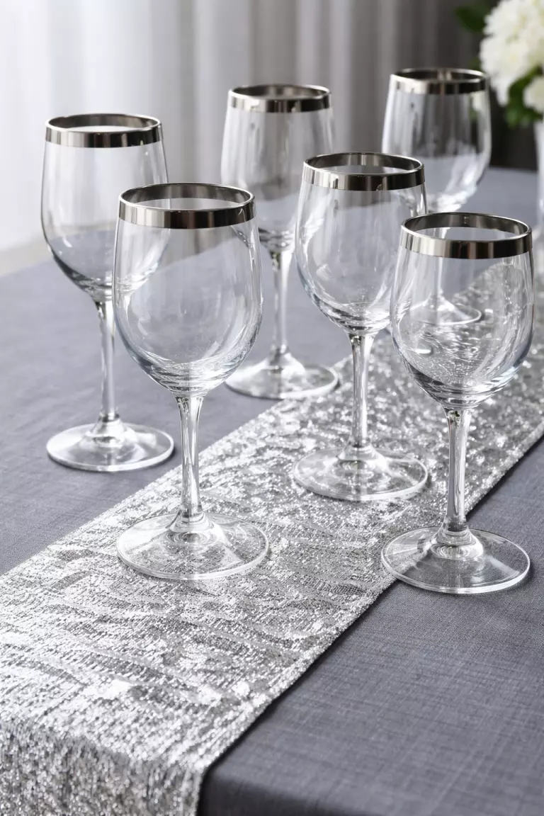A realistic photo of several silver rimmed wine glasses and a silver foil table runner on a grey tablecloth.