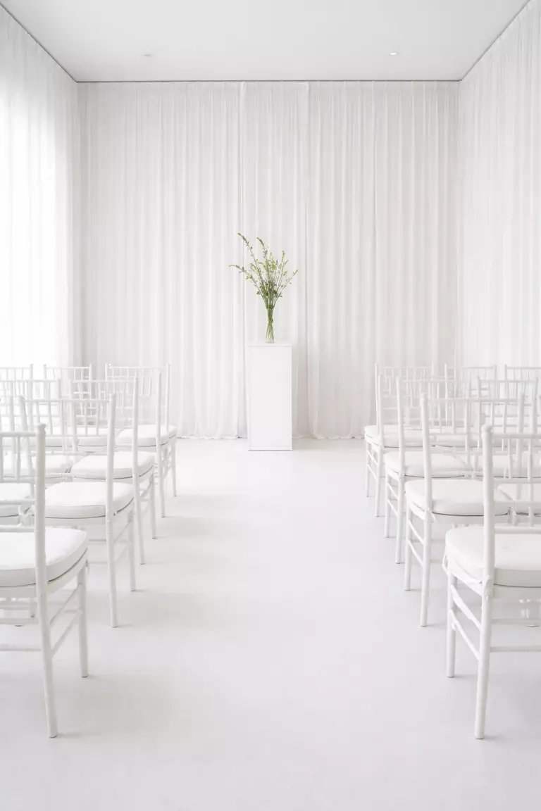 A realistic photo of a minimalist wedding ceremony room with all-white chairs, white floor-to-ceiling drapes, and a single geometric white pedestal holding a few green stems.