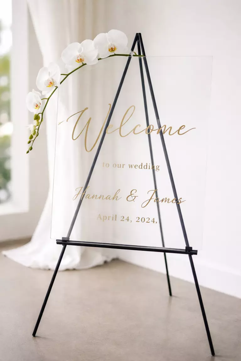 A realistic photo of a clear acrylic welcome sign with elegant gold calligraphy, resting on a minimalist black metal easel and decorated with a single long stem white orchid.