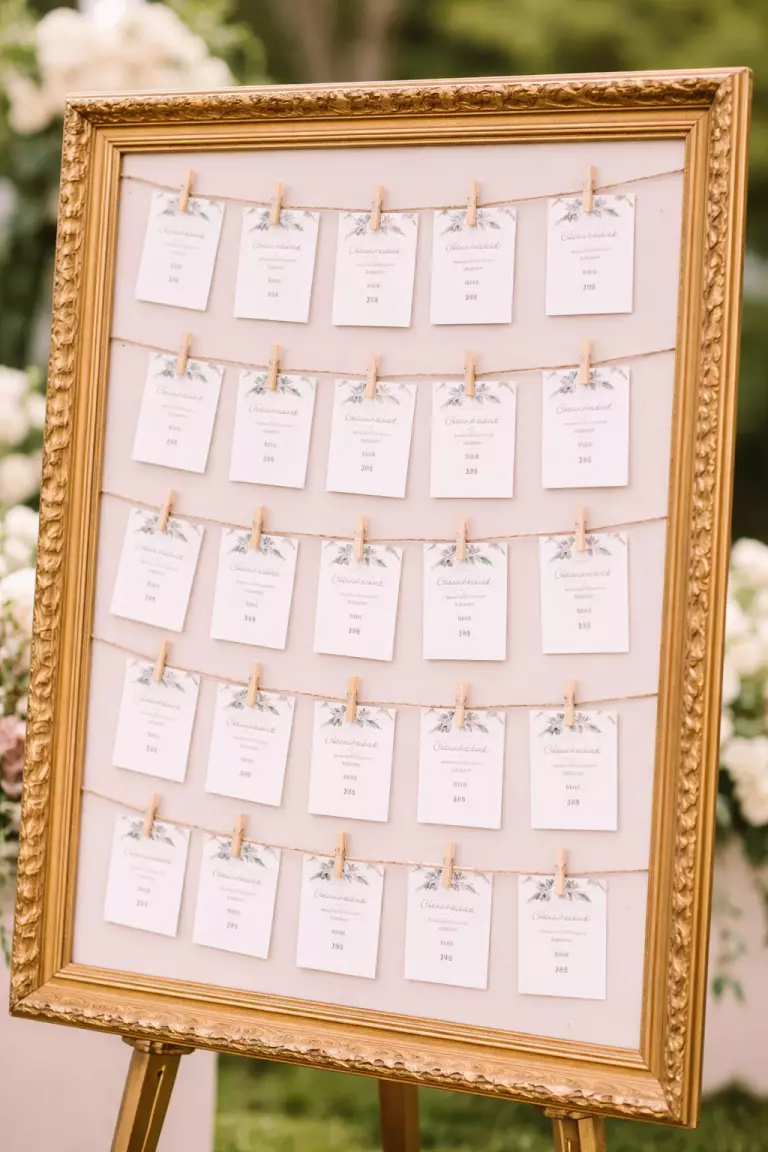 A realistic photo of a large gold frame with rows of string and small wooden clothespins holding photo cards with guest names and table numbers.