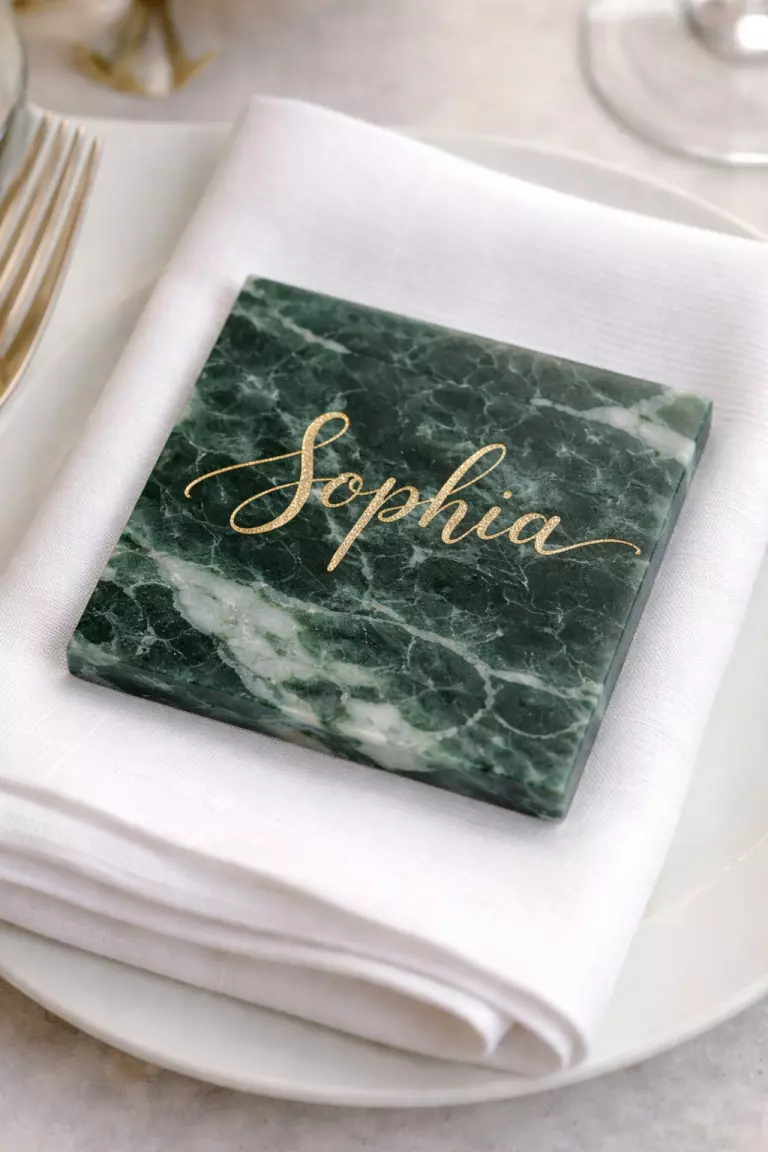 A realistic photo of a natural green marble tile with a guest name written in gold ink sitting on a white linen napkin.
