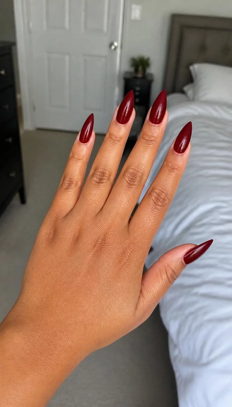Deep Crimson Stiletto Edge A photorealistic close-up shot of a woman's tan skin tone hand with five fingers, showcasing pointed long nails featuring a striking deep crimson color, bedroom background