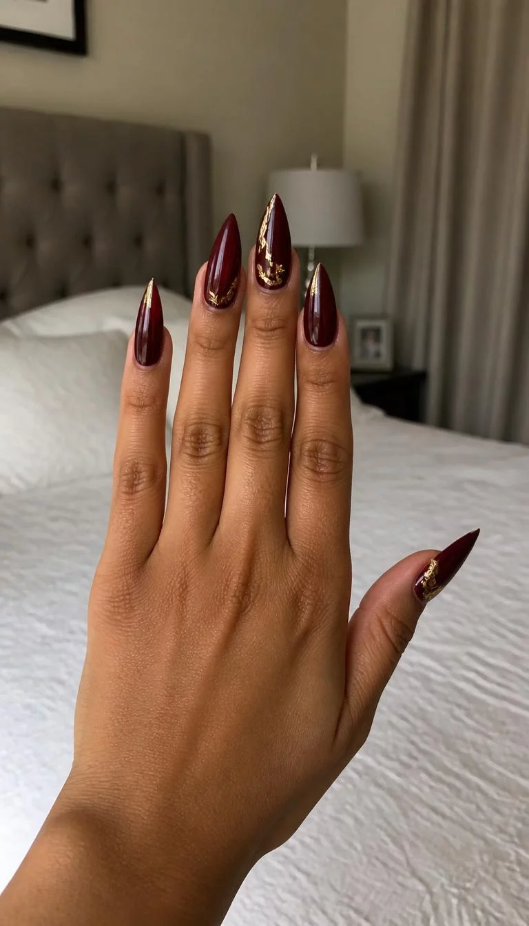 A photorealistic close-up shot of a woman's olive skin tone hand with five fingers, showcasing stiletto-shaped long nails in a deep burgundy, with a gold leaf accent design, bedroom background