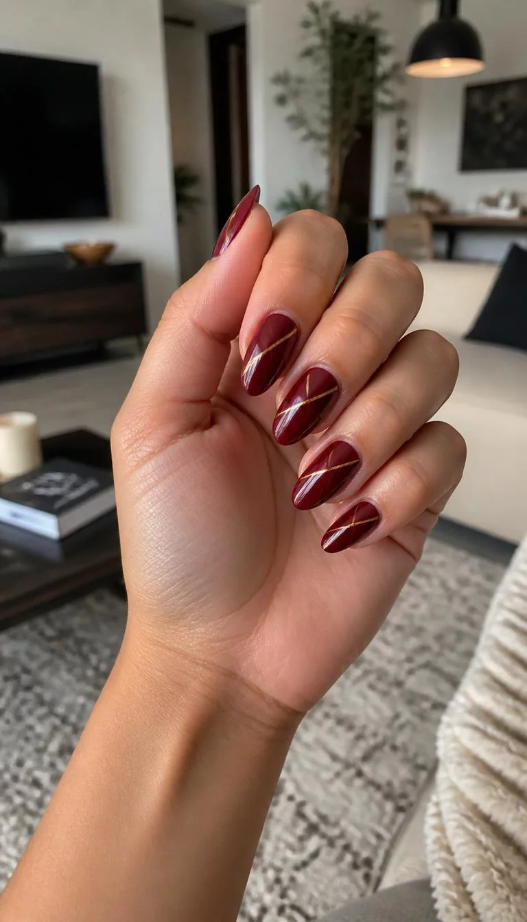 A photorealistic close-up shot of a woman's olive skin tone hand with five fingers, showcasing almond-shaped medium-length nails featuring a deep burgundy, accented with diagonal gold stripes, living room background