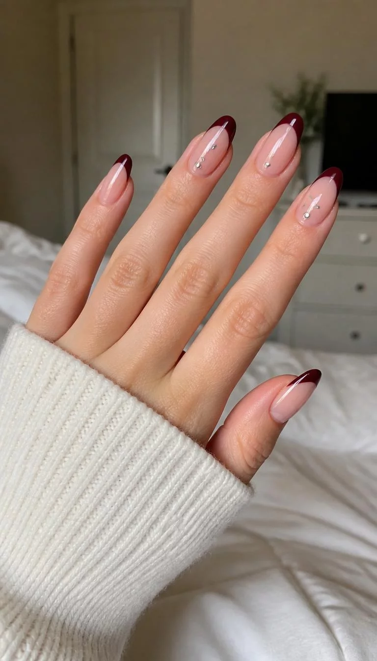 A photorealistic close-up shot of a woman's light skin tone hand with five fingers, showcasing almond-shaped medium-length nails featuring a classic French tip in deep burgundy and subtle nail gems, bedroom background