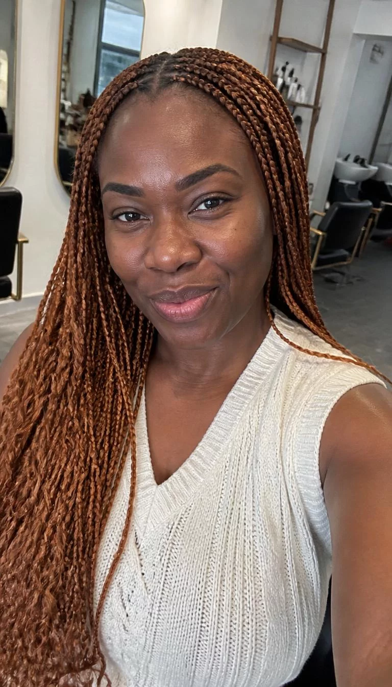 Copper Tinted Strawberry Blonde selfie of a 35-year-old Black African woman with a round face, dark brown, aquiline nose, thick natural eyebrows, and thin lips. Her face features slightly crooked smile, visible pores, and subtle natural facial asymmetry. She has monolid dark brown eyes, high cheekbones, broad smile, playful. She is athletic and is wearing a White Sweater Vest with copper strawberry blonde long braided hair, soft window light, hair salon