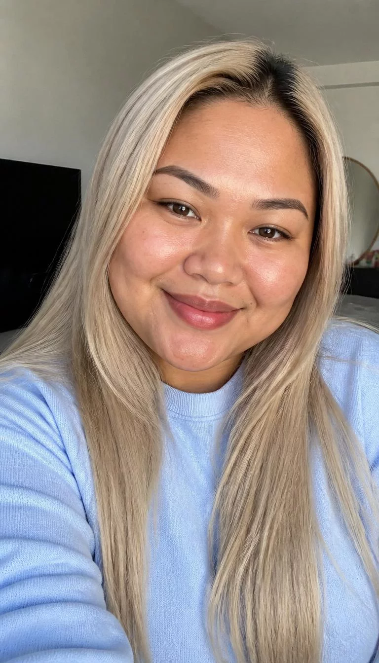 Pearlescent Almond Blonde selfie of a 31-year-old Southeast Asian woman with a square face, golden brown, button nose, thin arched eyebrows, and full lips. Her face features beauty mark near the lip, subtle acne marks, and subtle natural facial asymmetry. She has downturned brown eyes, high cheekbones, broad smile, confident. She is plus-size and is wearing a Light Blue Crewneck Sweatshirt with pearlescent almond blonde long straight hair, bright natural daylight, indoor