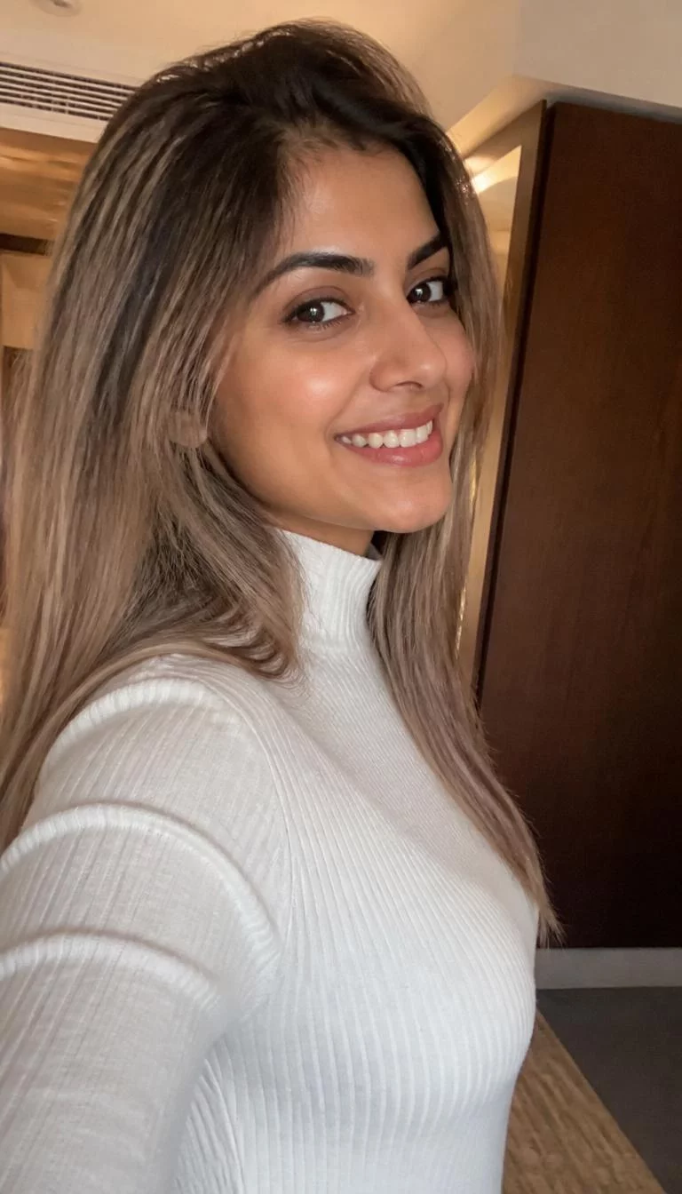 Smoky Mushroom Blonde Ombre selfie A side photo of a 27-year-old South Asian woman with a diamond face, deep brown, button nose, straight eyebrows, and heart-shaped lips. Her face features widow's peak hairline, a few laugh lines, and subtle natural facial asymmetry. She has upturned dark brown eyes, pronounced cheekbones, broad smile, cheerful. She is average and is wearing a White Mock Neck Sweater with smoky mushroom blonde long straight hair, warm indoor lighting, indoor