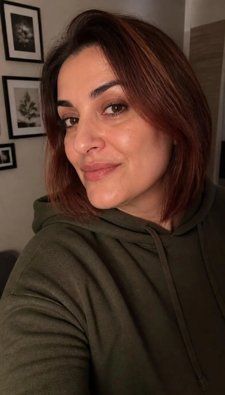Burnt Sienna Ombre A side photo selfie of a 40-year-old Southern European woman with a oval face, light, narrow nose, thick natural eyebrows, and full lips. Her face features slightly crooked smile, faint under-eye shadows, and subtle natural facial asymmetry. She has round dark brown eyes, pronounced cheekbones, closed-mouth smile, thoughtful. She is plus-size and is wearing a Olive Green Hoodie with burnt sienna reddish-brown ombre graduated bob hair, warm indoor lighting, indoor