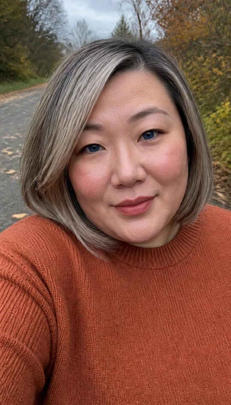 Smoky Quartz Blonde selfie of a 35-year-old East Asian woman with a oblong face, fair, aquiline nose, softly curved eyebrows, and full lips. Her face features slightly crooked smile, slightly flushed cheeks, and subtle natural facial asymmetry. She has monolid blue eyes, soft cheekbones, closed-mouth smile, thoughtful. She is plus-size and is wearing a Burnt Orange Sweater with smoky quartz ash blonde and gray bob hair, overcast daylight, fall outdoor background