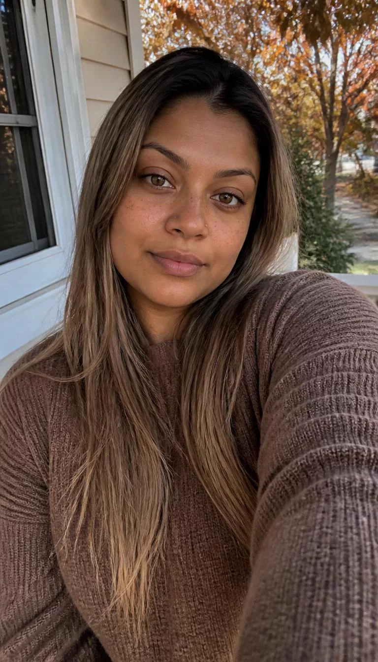 Sandy Driftwood Brown selfie of a 26-year-old South Asian woman with a square face, golden brown, broad nose, thin arched eyebrows, and wide lips. Her face features widow's peak hairline, light freckles, and subtle natural facial asymmetry. She has upturned hazel eyes, rounded cheeks, neutral expression, relaxed. She is curvy and is wearing a Brown Oversized Sweater with light ash brown with beige highlights long straight hair, soft window light, fall outdoor background