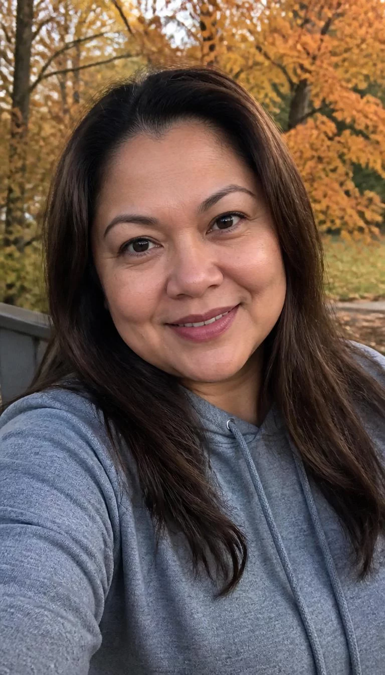 Velvet Truffle selfie of a 49-year-old Southeast Asian woman with a pear-shaped face, deep brown, button nose, thin arched eyebrows, and heart-shaped lips. Her face features widow's peak hairline, faint under-eye shadows, and subtle natural facial asymmetry. She has round dark brown eyes, pronounced cheekbones, broad smile, relaxed. She is plus-size and is wearing a Gray Hoodie with neutral dark matte brown long hair, soft window light, fall outdoor background