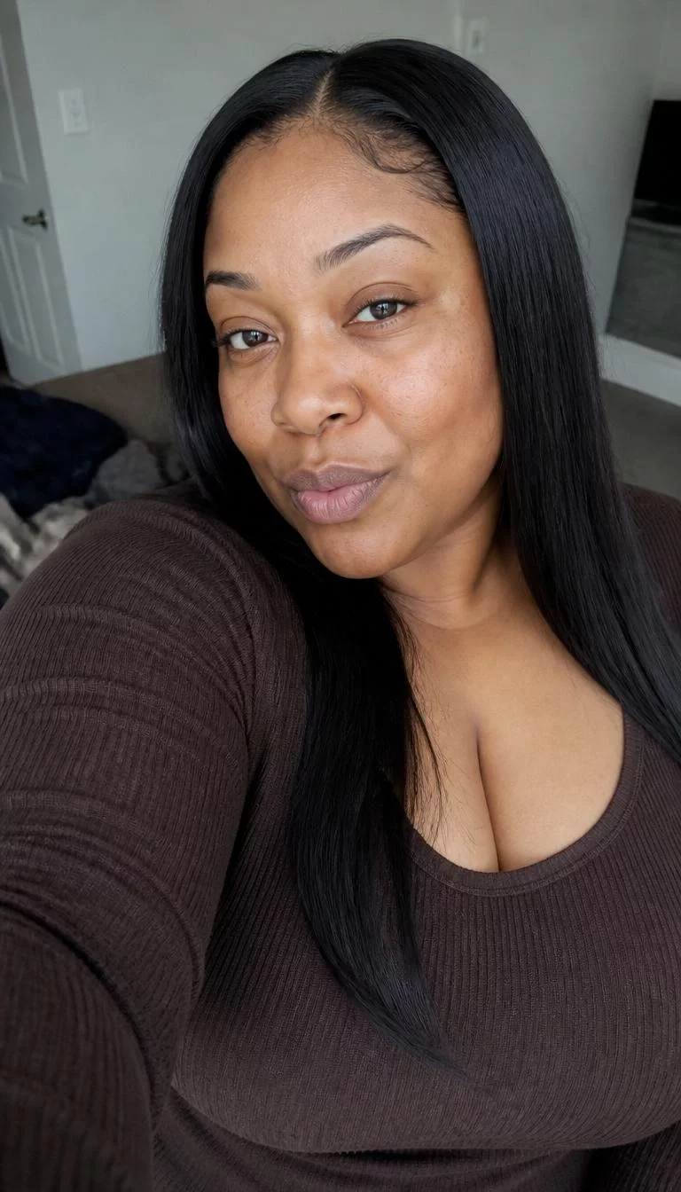 Espresso Bean Gloss selfie of a 41-year-old Black woman with a heart-shaped face, dark brown, upturned nose, feathered eyebrows, and full lips. Her face features uneven eyebrows, light freckles, and subtle natural facial asymmetry. She has monolid brown eyes, pronounced cheekbones, neutral expression, playful. She is plus-size and is wearing a Chocolate Brown Ribbed Top with dark espresso black long straight hair, overcast daylight, indoor