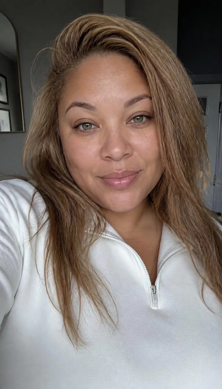 selfie of a 43-year-old Mixed-race woman with a heart-shaped face, dark brown, button nose, straight eyebrows, and wide lips. Her face features faint freckles across the nose, subtle acne marks, and subtle natural facial asymmetry. She has monolid green eyes, rounded cheeks, subtle smile, confident. She is plus-size and is wearing a white quarter-zip pullover with golden toffee blonde long textured hair, overcast daylight, indoor