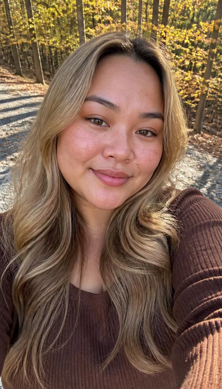 Cool Sand Dune selfie of a 29-year-old Southeast Asian woman with a oval face, light, broad nose, feathered eyebrows, and wide lips. Her face features uneven eyebrows, light freckles, and subtle natural facial asymmetry. She has downturned hazel eyes, high cheekbones, closed-mouth smile, cheerful. She is plus-size and is wearing a Brown Knit Top with sandy blonde long wavy hair, bright natural daylight, fall outdoor background