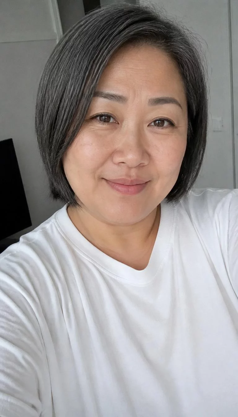 Soft Charcoal Smoke selfie of a 44-year-old East Asian woman with a round face, deep brown, narrow nose, thick natural eyebrows, and full lips. Her face features slightly crooked smile, a few laugh lines, and subtle natural facial asymmetry. She has almond-shaped brown eyes, rounded cheeks, closed-mouth smile, thoughtful. She is plus-size and is wearing a white oversized t-shirt with muted ash gray chin length bob hair, overcast daylight, indoor