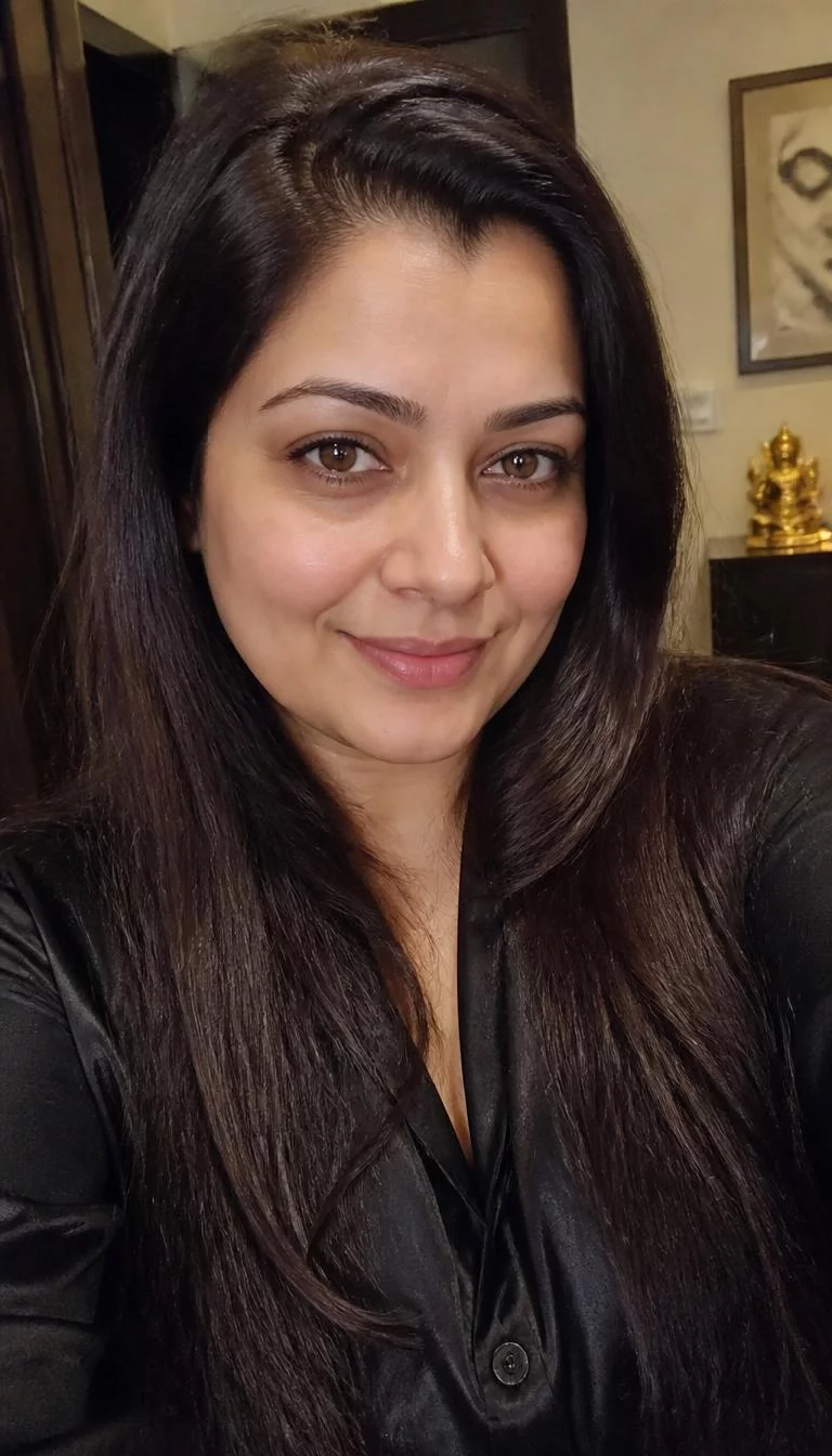 Deep Roasted Espresso selfie of a 44-year-old South Asian woman with a heart-shaped face, fair, straight nose, thin arched eyebrows, and heart-shaped lips. Her face features widow's peak hairline, faint under-eye shadows, and subtle natural facial asymmetry. She has round brown eyes, rounded cheeks, closed-mouth smile, relaxed. She is plus-size and is wearing a black silk blouse with deep roasted espresso long sleek straight hair, warm indoor lighting, indoor