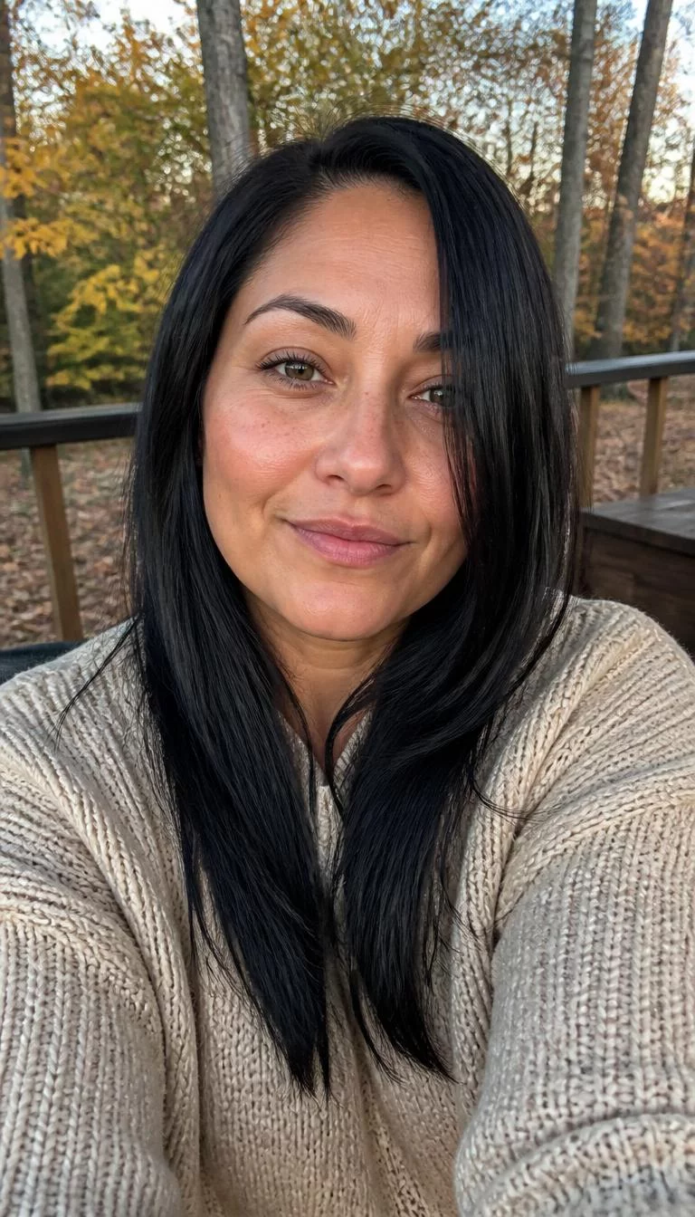 Midnight Navy Tint selfie of a 39-year-old Latina woman with a oblong face, light, broad nose, softly curved eyebrows, and heart-shaped lips. Her face features faint freckles across the nose, light freckles, and subtle natural facial asymmetry. She has hooded hazel eyes, pronounced cheekbones, subtle smile, thoughtful. She is athletic and is wearing a beige chunky knit sweater with midnight navy tint long blunt cut hair, soft window light, fall outdoor background