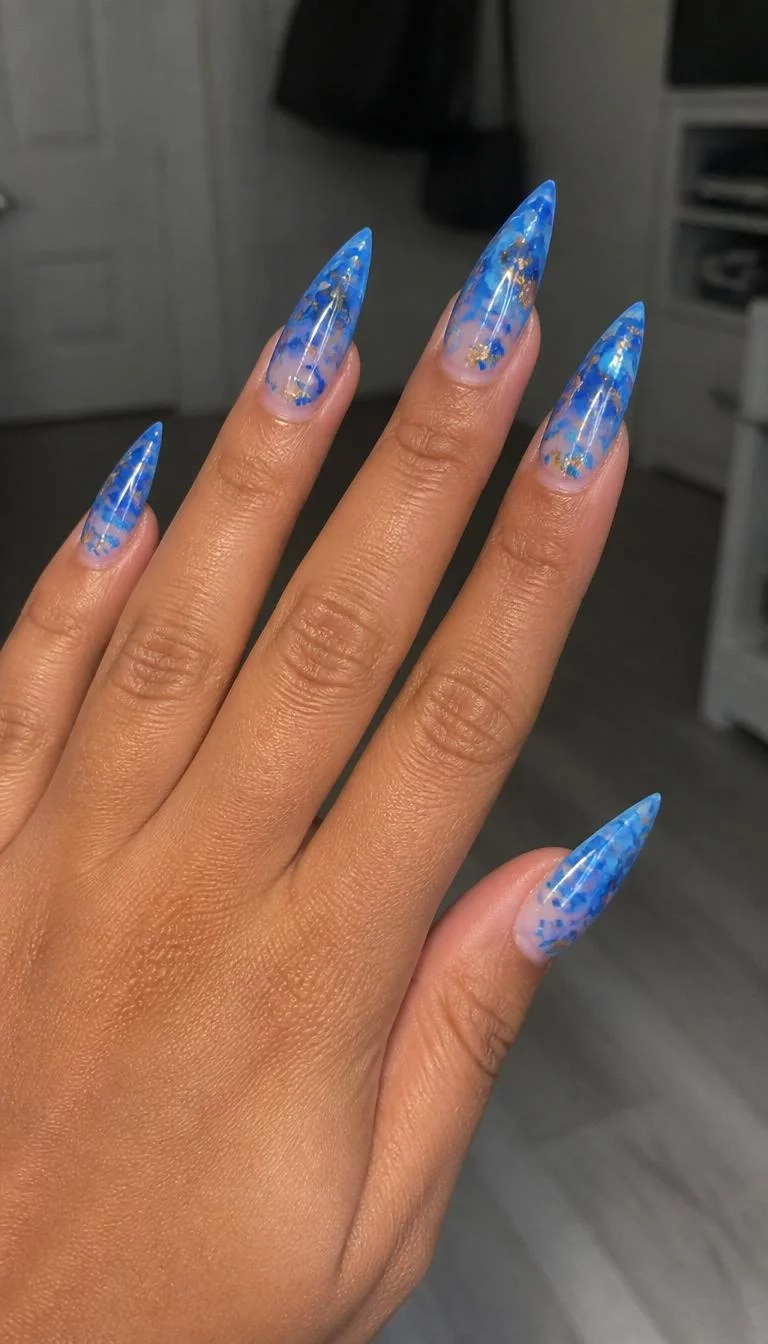 Blue Marble Stiletto A photorealistic close-up shot of a woman's medium skin tone hand with five fingers, showcasing long stiletto-shaped nails featuring a bright blue color and a sheer, marble-like design with rose gold flakes, indoor background