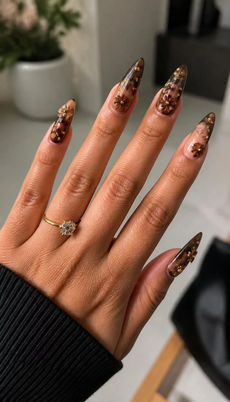 Dark Brown 3D Florals A photorealistic close-up shot of a woman's medium skin tone hand with five fingers, showcasing long pointed nails featuring a sophisticated design with 3D floral accents, a dark brown gradient, and gold studs, indoor background