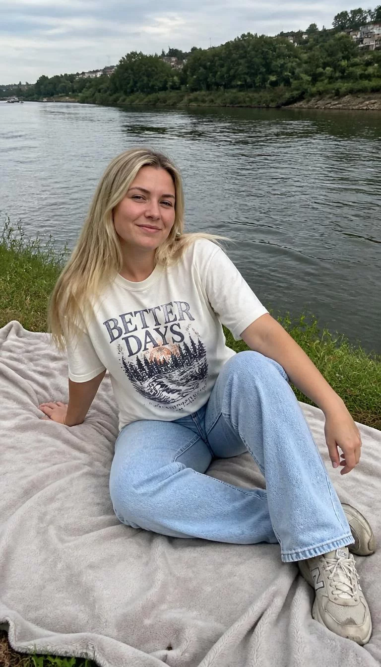 Riverfront Casual in Denim and Tees A photorealistic full body shot of a 29-year-old Northern European woman with a round face, olive, upturned nose, thick natural eyebrows, and thin lips. Her face features slightly crooked smile, a few laugh lines, and subtle natural facial asymmetry. She has upturned hazel eyes, rounded cheeks, closed-mouth smile, playful. She is athletic build with blonde straight hair and is wearing a white graphic t-shirt, light wash denim jeans, sneakers, sitting on a blanket by a river, everyday social media post aesthetic, high-quality mobile photography, shot on an iPhone 14.
