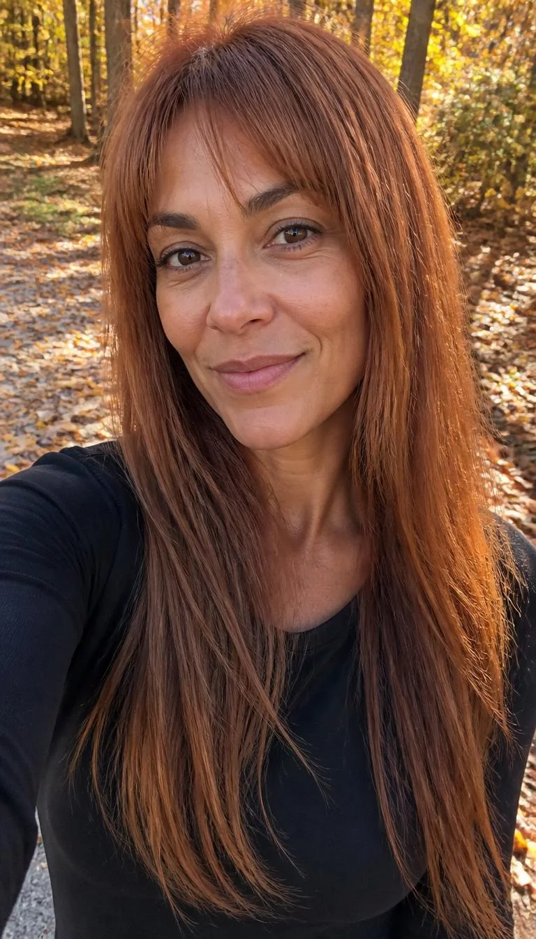 Natural Terra Cotta selfie of a 49-year-old Mixed-race woman with a heart-shaped face, fair, narrow nose, thick natural eyebrows, and heart-shaped lips. Her face features small scar on the eyebrow, faint under-eye shadows, and subtle natural facial asymmetry. She has deep-set brown eyes, pronounced cheekbones, closed-mouth smile, playful. She is athletic and is wearing a Black Long Sleeve Tee with Natural Terra Cotta long straight with fringe hair, bright natural daylight, fall outdoor background