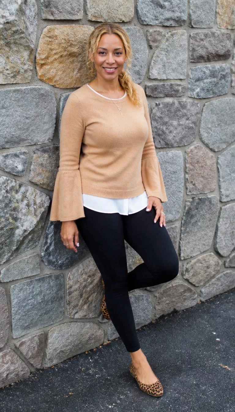 A photorealistic full body shot of a 46-year-old Mixed-race woman with a oblong face, light, broad nose, softly curved eyebrows, and thin lips. Her face features slightly crooked smile, faint under-eye shadows, and subtle natural facial asymmetry. She has deep-set dark brown eyes, pronounced cheekbones, broad smile, cheerful. She is slim with blonde hair pulled back in loose waves and is wearing a tan sweater with bell sleeves over a white blouse, black leggings, and leopard print flats, posing against a stone wall, everyday social media post aesthetic, high-quality mobile photography, shot on an iPhone 14.