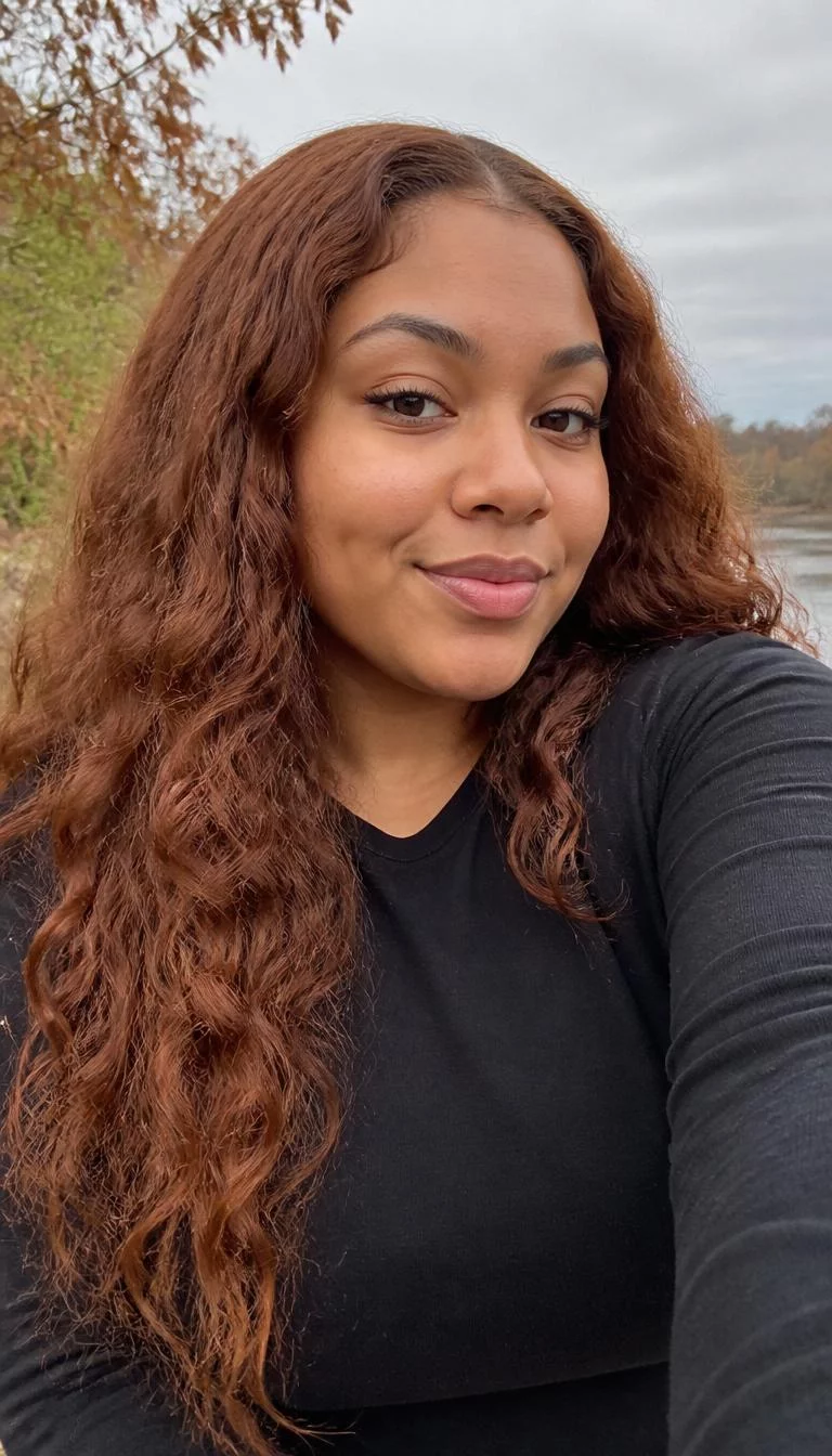 Burnt Sienna Melt selfie of a 23-year-old Mixed-race woman with a oblong face, golden brown, button nose, feathered eyebrows, and full lips. Her face features dimples, subtle acne marks, and subtle natural facial asymmetry. She has upturned amber eyes, soft cheekbones, closed-mouth smile, confident. She is plus-size and is wearing a Black Long Sleeve Tee with burnt sienna melt long crimped waves hair, overcast daylight, fall outdoor background