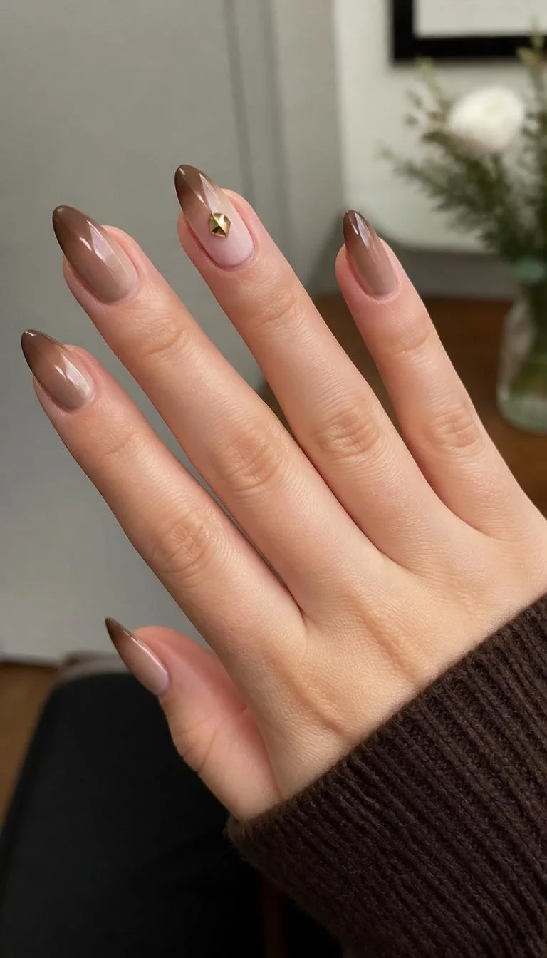 Coffee Bean Gradient A photorealistic close-up shot of a woman's fair skin tone hand with five fingers, showcasing almond-shaped medium-length nails featuring a gradient of light cream to dark brown, accented with a golden gem, indoor background